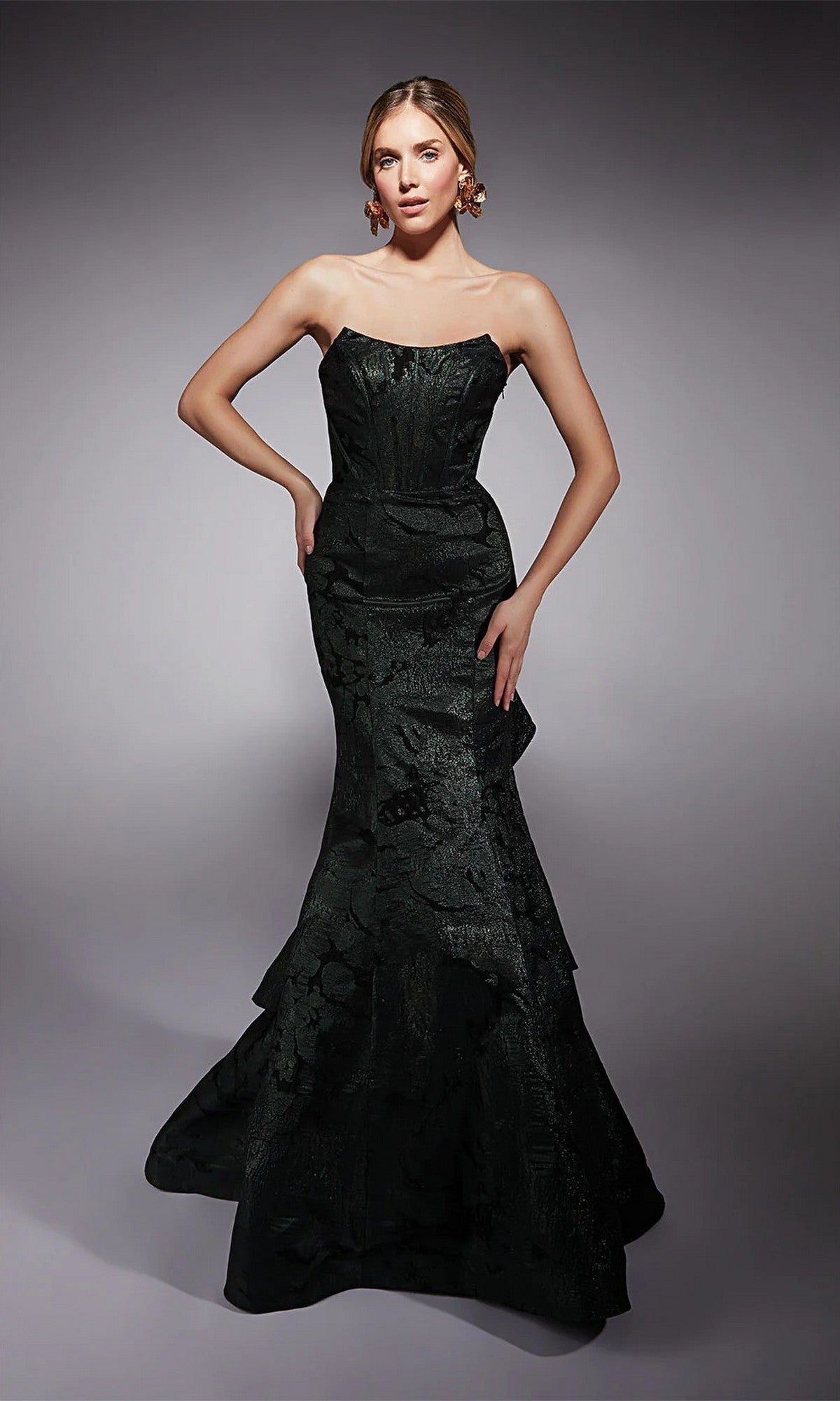 A woman poses against a gray background in the Alyce 27747 long formal dress, featuring a strapless fitted bodice and flared mermaid skirt crafted from textured black fabric.